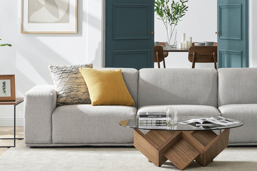 A living room with a gray couch and yellow pillows with a table in front of it made of wood and glass 