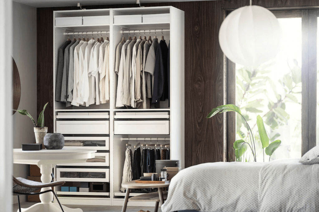 A white closet with clothes hanging in it and a white bed in front of a window 